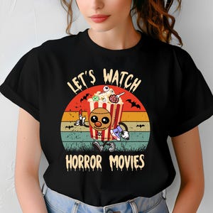 Peut inclure: T-shirt noir avec un graphisme coloré représentant un seau de pop-corn avec un visage cousu, levant un doigt et une hache. Le texte dit "Let's Watch Horror Movies" dans un style vintage.