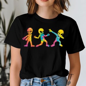 May include: Black t-shirt with a graphic of three colourful cartoon aliens. The aliens are in different poses, including one dabbing. The aliens have large eyes and are outlined in black. The t-shirt is made of a soft material.