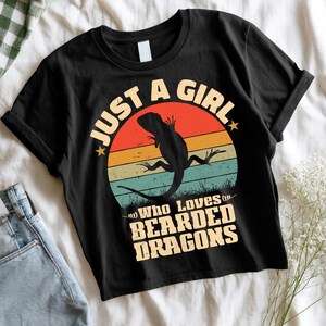 Peut inclure: T-shirt noir avec un motif circulaire représentant une silhouette de dragon barbu sur fond de coucher de soleil. Le texte indique "JUST A GIRL Who Loves BEARDED DRAGONS" dans un style vintage audacieux.