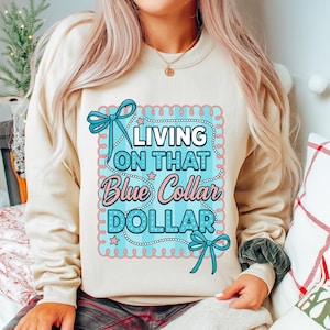 May include: A beige sweatshirt with the text "LIVING ON THAT Blue Collar DOLLAR" in a blue and pink design. The design includes bows, stars, and a scalloped border. The sweatshirt is a casual style.