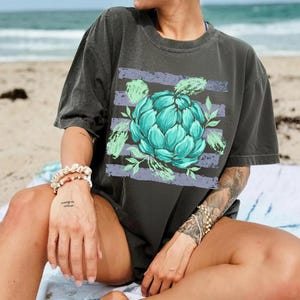 May include: Dark gray t-shirt featuring a teal artichoke design with green leaves and purple stripes. The graphic tee is a relaxed fit, perfect for casual wear. The shirt is made of a soft, comfortable material.
