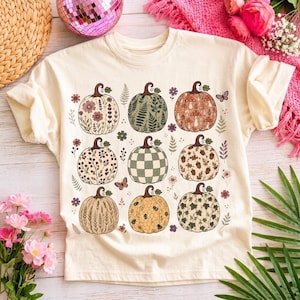 Peut inclure: Un t-shirt couleur crème arborant une impression graphique de neuf citrouilles disposées en grille. Chaque citrouille présente un motif unique, incluant des fleurs, fougères, champignons, carreaux, glands, blé, abeilles et lierre, avec des papillons.