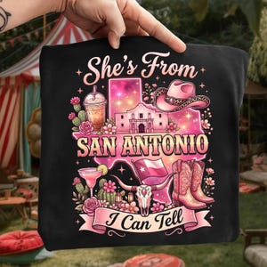 May include: Black graphic t-shirt with a pink and white design featuring the text "She's From San Antonio I Can Tell". The design includes a cowboy hat, boots, and the Alamo.