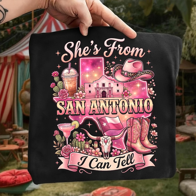 May include: Black graphic t-shirt with a pink and white design featuring the text "She's From San Antonio I Can Tell". The design includes a cowboy hat, boots, and the Alamo.