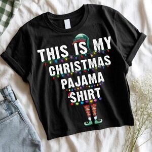 May include: Black t-shirt with the text "THIS IS MY CHRISTMAS PAJAMA SHIRT" in white, decorated with colorful Christmas lights and elf legs. The shirt has a rolled sleeve and a green elf hat graphic.