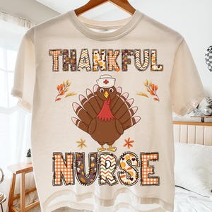 Peut inclure: T-shirt crème avec l'inscription "THANKFUL NURSE" dans un motif patchwork. Une dinde de dessin animé portant un chapeau d'infirmière est centrée sous le mot "THANKFUL". La dinde a des plumes marron, un fanon rouge et un chapeau blanc avec une croix rouge.