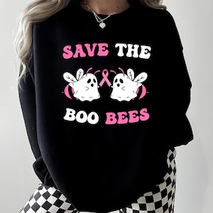 Puede incluir: Sudadera negra con el texto "SAVE THE BOO BEES" en blanco y rosa. El diseño presenta dos fantasmas de dibujos animados con alas y una cinta rosa, que simboliza la concienciación sobre el cáncer de mama.