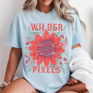 以下が含まれることがあります： 大きな赤い花のグラフィックと「WILDER PIXELS」の文字が赤色でプリントされた、水色のTシャツ。花は紫色の中心を持ち、緑の葉と小さな赤い花に囲まれています。カジュアルなスタイルのシャツです。