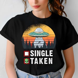 Puede incluir: Camiseta negra con un diseño retro de un OVNI sobre un atardecer y árboles. El texto dice "SINGLE" con una casilla de verificación en blanco y "TAKEN" con una casilla de verificación con una cabeza de alienígena. Una camiseta divertida y novedosa.