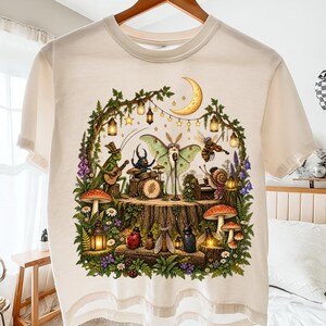 Peut inclure: Un t-shirt à manches courtes de couleur crème avec une impression graphique d'insectes jouant de la musique sur une souche d'arbre. La scène montre une sauterelle jouant de la guitare, un scarabée à la batterie, un papillon chantant et un escargot au piano.