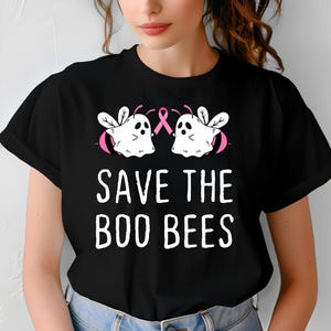 Puede incluir: Camiseta negra con el texto blanco "SAVE THE BOO BEES". El diseño incluye dos abejas blancas con forma de fantasma con detalles rosas y una cinta rosa. Una prenda informal.