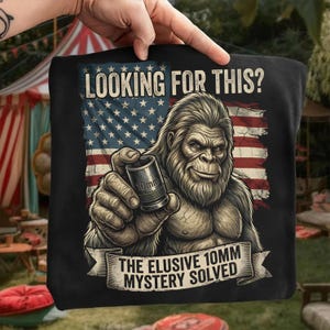 Könnte beinhalten: Schwarzes T-Shirt mit einer Grafik von Bigfoot, der einen 10-mm-Steckschlüssel vor einer amerikanischen Flagge hält. Der Text lautet "LOOKING FOR THIS?" und "THE ELUSIVE 10MM MYSTERY SOLVED" auf einem Banner.