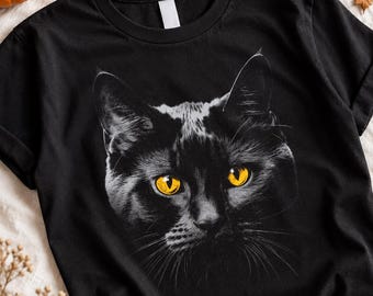 May include: A black short-sleeved t-shirt featuring a detailed graphic print of a cat face. The cat is rendered in shades of black and white with striking, bright yellow eyes that stand out against the dark fur and the black fabric of the shirt.