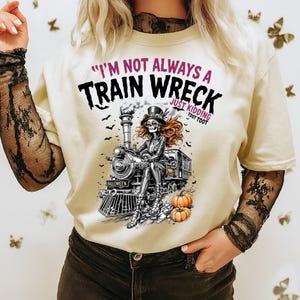 Puede incluir: Camiseta color crema con un diseño de Halloween. El diseño presenta una mujer esqueleto en un tren, calabazas y murciélagos. El texto dice "I'm Not Always a Train Wreck Just Kidding Toot Toot".