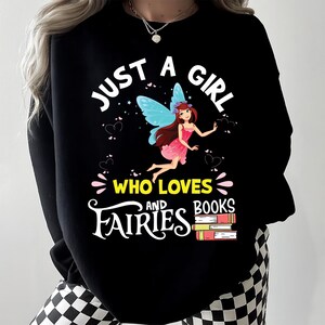 Peut inclure: Sweat-shirt noir avec l'inscription "JUST A GIRL WHO LOVES FAIRIES AND BOOKS". Le motif représente une fée aux ailes bleues et une robe rose, avec des cœurs et une pile de livres. Le sweat-shirt est de style décontracté.