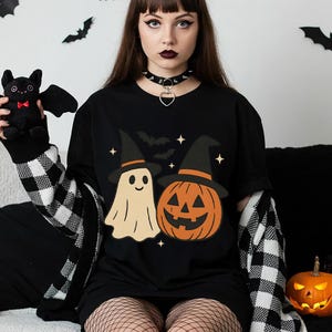 Peut inclure: Un t-shirt noir arborant une impression graphique représentant un fantôme crème et une citrouille d'Halloween orange, tous deux coiffés de chapeaux de sorcière noirs. Le motif inclut des étoiles et des silhouettes de chauves-souris volantes.