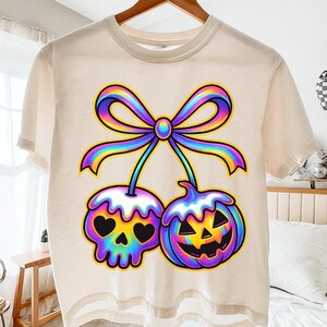 Peut inclure: T-shirt crème avec un motif coloré de deux motifs stylisés sur le thème d'Halloween : un crâne et une citrouille, reliés par un nœud arc-en-ciel. Le crâne a des yeux en forme de cœur. Le motif est souligné en noir.