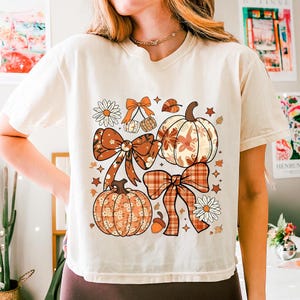 Peut inclure: T-shirt crème avec un motif sur le thème de l'automne. Le design comprend des citrouilles, des nœuds, des feuilles et des marguerites dans des tons orange, marron et crème. Le t-shirt a une coupe décontractée et des manches courtes.