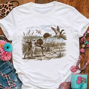 Op de afbeelding: Wit t-shirt met een gedetailleerde illustratie van een jachthond in een moerasomgeving. De hond is bruin en wit, met eenden op de achtergrond. Het ontwerp heeft een vintage, outdoor esthetiek, geschikt voor liefhebbers van natuur en jacht.