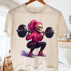 May include: Cream-colored t-shirt featuring a cartoon skeleton with pink hair lifting a barbell. The skeleton is wearing a pink hoodie, black leggings, and pink sneakers. The design has a fitness theme.
