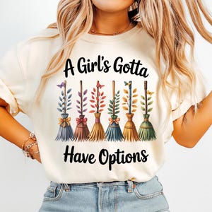 May include: A cream-colored t-shirt featuring the black text A Girl's Gotta Have Options. Below the text are six stylized broomsticks in various colors, including blue, brown, yellow, and green, each adorned with small leaves and butterflies.