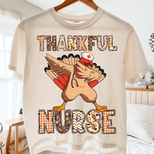 Peut inclure: T-shirt beige avec une dinde de dessin animé en uniforme d'infirmière, en train de faire un dab. Le texte "THANKFUL NURSE" est imprimé au-dessus et en dessous de la dinde dans une police sur le thème de l'automne. La dinde porte une coiffe d'infirmière et un stéthoscope.