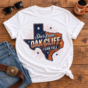 Peut inclure: Un t-shirt blanc à manches courtes avec un motif représentant l'État du Texas en bleu marine et orange. Le design inclut le texte She's from OAK CLIFF I CAN TELL, entouré de petites étoiles et de lignes rayonnantes dans un style vintage usé.