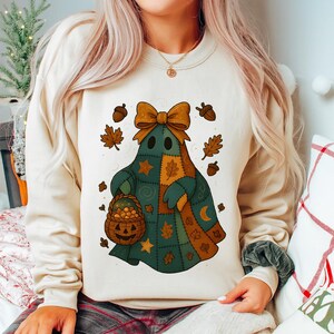 Peut inclure: Un sweat-shirt crème avec un motif de fantôme patchwork. Le fantôme est orné d'un grand nœud, de feuilles d'automne, de glands et d'un panier citrouille. Le design utilise des nuances de vert, d'orange et de marron, parfait pour l'automne.