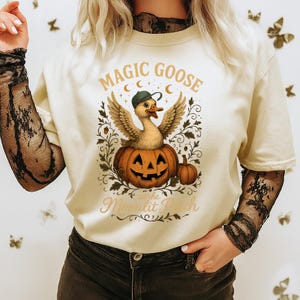Puede incluir: Camiseta color crema con una ilustración caprichosa de un ganso con gorra, emergiendo de una calabaza tallada. El texto "MAGIC GOOSE at the Moonlit Patch" está arqueado arriba y abajo de la imagen, en un estilo vintage.