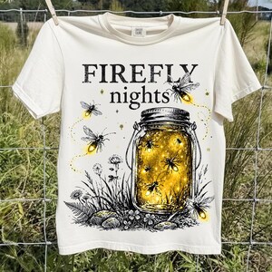 Peut inclure: Un t-shirt couleur crème avec une impression graphique représentant un bocal en verre rempli de lucioles jaunes lumineuses. Le texte "FIREFLY nights" est imprimé au-dessus du bocal, entouré d'illustrations de lucioles et de fleurs sauvages.