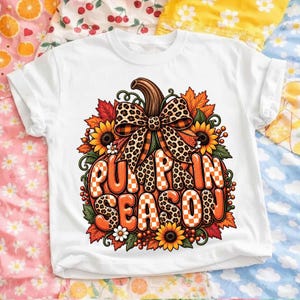 Peut inclure: Un t-shirt blanc avec un motif représentant une grosse citrouille orange ornée d'un nœud à imprimé léopard. Le texte PUMPKIN SEASON est inscrit sur la citrouille dans un mélange de motifs à carreaux orange et léopard, entouré de tournesols.