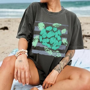 May include: Dark grey t-shirt featuring a large teal and green artichoke design with horizontal stripes. The shirt has a crew neck and short sleeves. The image is taken on a beach.