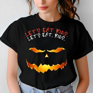 Op de afbeelding: Zwart T-shirt met de tekst "LET'S EAT KIDS" in rood en wit. Daaronder een pompoengezicht met oranje en gele accenten. Een Halloween-thema grafisch T-shirt.