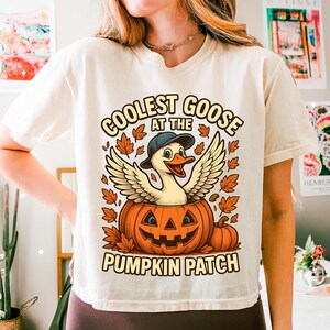 Peut inclure: T-shirt crème avec un dessin animé d'une oie portant une casquette et des ailes, sortant d'une citrouille sculptée. Le texte dit "COOLEST GOOSE AT THE PUMPKIN PATCH" dans un design sur le thème de l'automne.