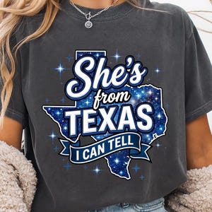 Peut inclure: Un t-shirt gris anthracite à manches courtes avec un motif de l'État du Texas rempli d'un imprimé galaxie bleu. Le texte indique She's from TEXAS I CAN TELL en lettres blanches et bleues, entouré de petits accents en forme d'étoiles.