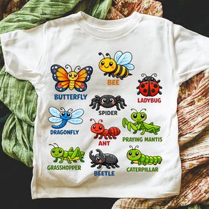 Peut inclure: T-shirt blanc avec des insectes de dessins animés colorés : papillon, abeille, coccinelle, libellule, araignée, fourmi, mante religieuse, sauterelle, scarabée et chenille. Chaque insecte a son nom imprimé en dessous.