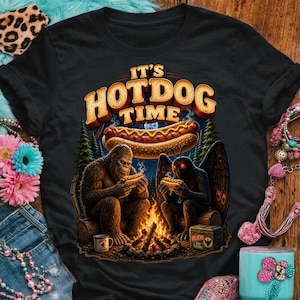 May include: Black t-shirt with a graphic featuring a Bigfoot and a Mothman enjoying hot dogs around a campfire. The text "IT'S HOTDOG TIME" is displayed above a floating hot dog.