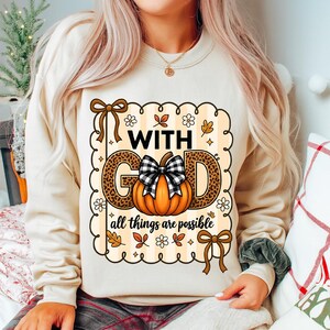 Peut inclure: Sweat-shirt beige avec un motif graphique portant l'inscription « WITH GOD all things are possible ». Le mot « GOD » est en imprimé léopard, avec une citrouille et un nœud à carreaux noir et blanc. Le motif comprend des feuilles d'automne et des fleurs.