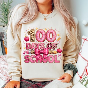 Puede incluir: Sudadera beige con un gráfico colorido que celebra los 100 días de escuela. El diseño incluye el número "100" en estilo donut, las palabras "Days of School", un lazo rosa, lápices y manzanas.