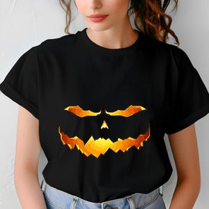 Op de afbeelding: Zwart T-shirt met een gloeiend pompoengezicht-ontwerp. De afbeelding heeft oranje en gele highlights, waardoor een griezelig Halloween-thema ontstaat. Het shirt is een klassieke ronde hals.