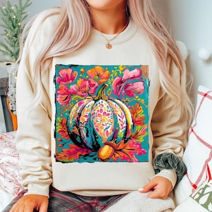 Peut inclure: Sweat-shirt crème avec un motif artistique vibrant. Il présente une citrouille turquoise ornée de motifs floraux colorés, entourée de fleurs roses et oranges et de feuilles d'automne. Un gland est également représenté. Vêtement d'automne confortable.