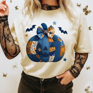Peut inclure: T-shirt crème avec un motif de citrouille patchwork bleu, un nœud floral, des étoiles et des silhouettes de chauves-souris. La citrouille a un visage sculpté. Un t-shirt graphique sur le thème d'Halloween.