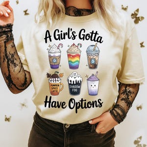 Peut inclure: Un t-shirt couleur crème avec le texte noir A Girl's Gotta Have Options et six illustrations de boissons. Les motifs incluent une boisson violette, une tasse aux rayures arc-en-ciel, un café, un latte à la citrouille, une boisson shadow fog et un smoothie violet.