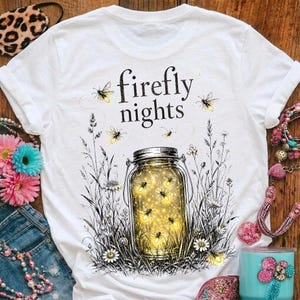 Peut inclure: Un t-shirt blanc avec un motif représentant un bocal en verre rempli de lucioles jaunes lumineuses. Le texte "firefly nights" est imprimé au-dessus du bocal dans une police noire. Le dessin inclut des herbes hautes et des fleurs sauvages.