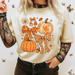 Peut inclure: T-shirt crème avec un motif sur le thème de l'automne. Le graphisme comprend des citrouilles oranges, des nœuds, des fleurs et des étoiles. Les citrouilles ont des motifs floraux et les nœuds ont un imprimé léopard. Vêtement décontracté et saisonnier.