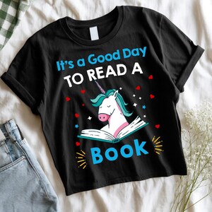 Peut inclure: T-shirt noir avec le texte "It's a Good Day to Read a Book". Le t-shirt présente une licorne lisant un livre, avec des cœurs et des étoiles. La licorne a une crinière et une corne bleues.