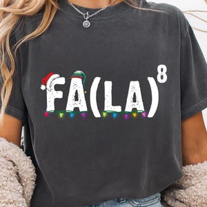 May include: Dark gray t-shirt with the text "FA(LA)8" in white. The letters "FA" have a Santa hat, and the "LA" has a green elf hat. The text is above a string of colorful Christmas lights. A festive holiday tee.