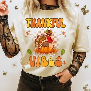 Peut inclure: T-shirt crème avec les mots "THANKFUL VIBES" en orange et jaune. Une dinde de dessin animé portant un chapeau de pèlerin est assise sur des citrouilles. Des feuilles d'automne et des motifs floraux entourent la dinde.