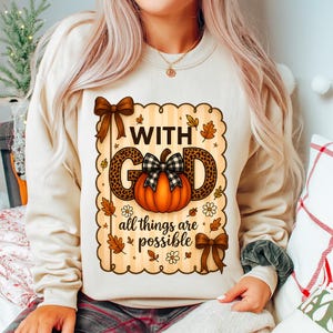 Peut inclure: Sweat-shirt beige avec un motif sur le thème de l'automne. Le motif comprend les mots "WITH GOD" au-dessus d'une citrouille avec un nœud à carreaux, et la phrase "all things are possible" en dessous. Des nœuds bruns et des feuilles d'automne complètent le design.