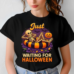 Peut inclure: T-shirt noir avec un graphisme coloré d'Halloween. Le motif représente un squelette se relaxant dans une bouée avec une citrouille et le texte "Just Waiting for Halloween".